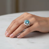swiss blue topaz round stone 29.50ct  Hand wearing a ring with a blue gemstone on a marble surface