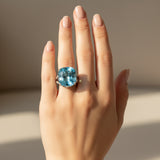 swiss blue topaz round stone 39.85ct  Hand wearing a ring with a blue gemstone on a neutral background