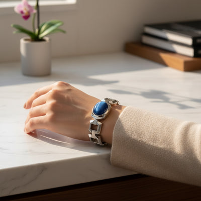 cats eye stone 57.35ct Hand wearing a silver bracelet with a blue stone on a light surface, with a plant and books in the background.
